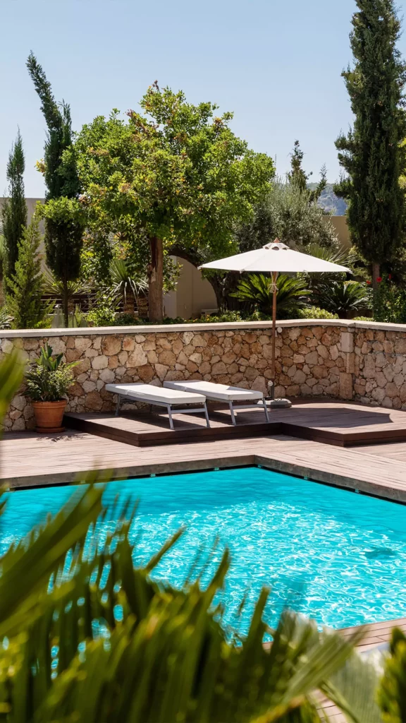 Pool mit zwei Liegen und Sonnenschirm auf Holzdeck, umgeben von mediterranen Pflanzen und Steinmauer bei klarem Himmel